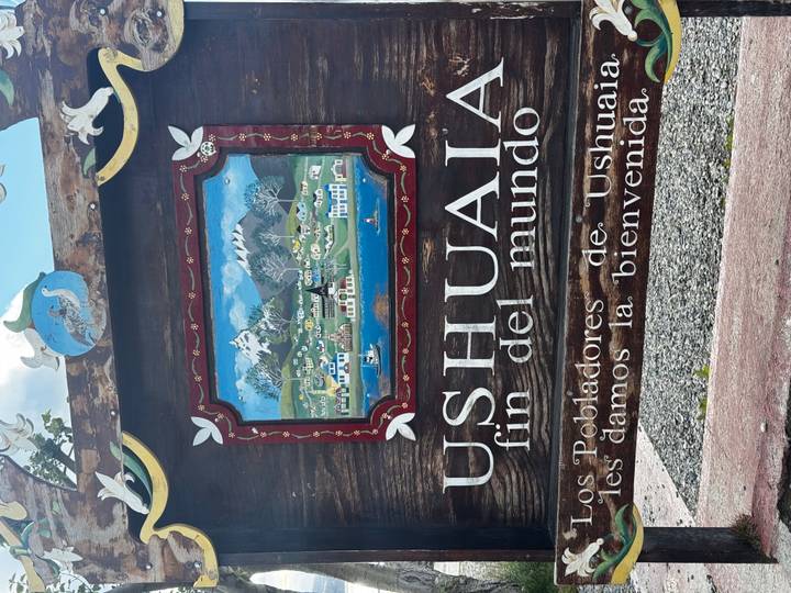 Painted wooden welcome sign reading "USHUAIA fin del mundo" decorated with a colourful town illustration.