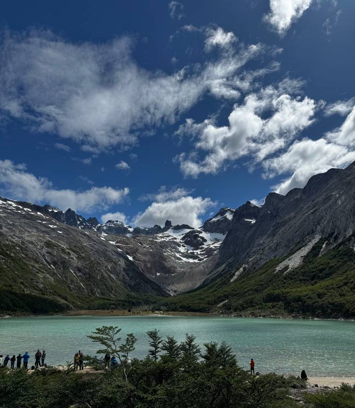 Wide valley surrounded by dramatic craggy peaks with patches of snow beneath a bright blue, cloud-dotted sky.