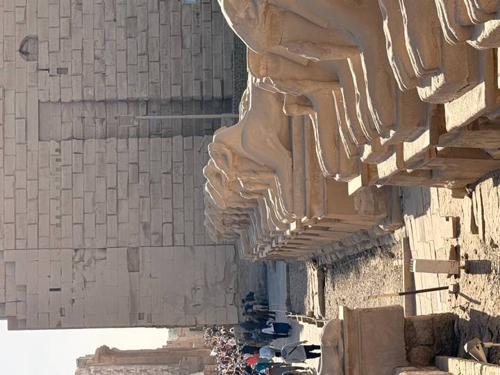 Row of ram-headed sphinx statues lining the entrance to Karnak Temple with visitors in the background.
