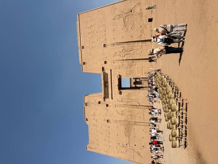 Large sandstone pylon of an ancient Egyptian temple with crowds and rows of empty chairs in the forecourt.