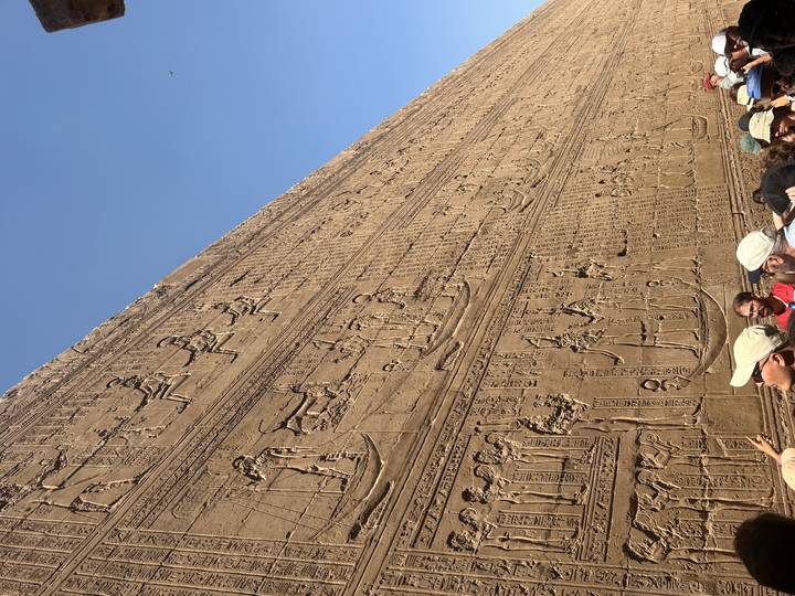 Massive sandstone wall densely carved with hieroglyphic reliefs viewed by tourists wearing hats.