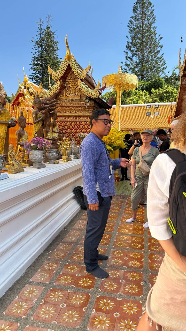 Tour guide explaining inside a Thai temple adorned with golden Buddha statues and floral offerings.