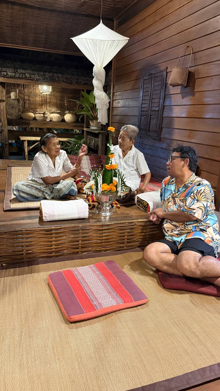 Two elderly Thai women and a guide seated on bamboo mats engaging in a cultural ceremony with flower arrangement.