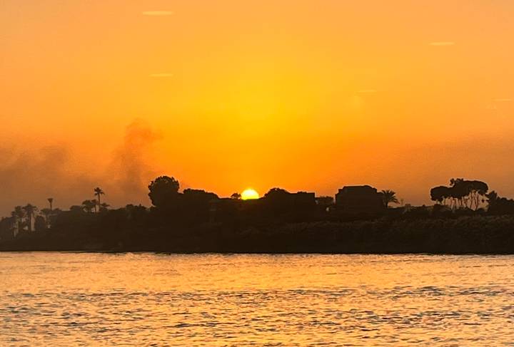 An intense orange sunset over the Nile river, silhouetting palm trees and low buildings with smoke rising in the distance.