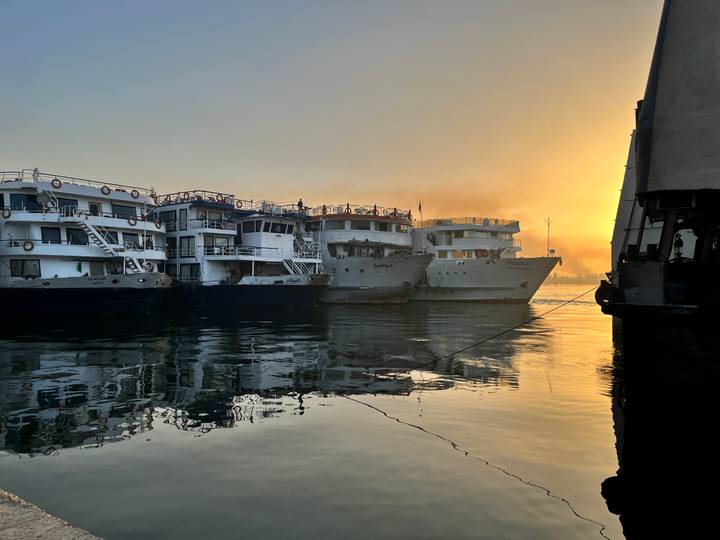 Several Nile cruise boats docked side by side at sunset, passengers visible on the top decks with golden light reflecting on calm water.