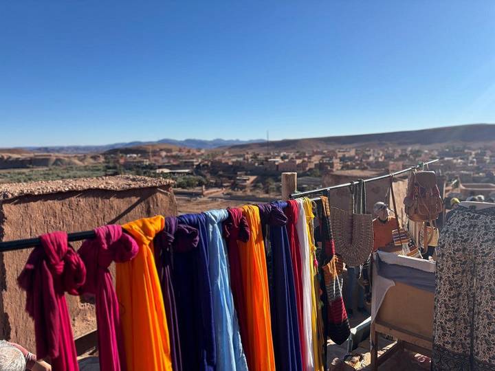Vivid fabric scarves hang drying above a desert village with distant ranges under blue sky.