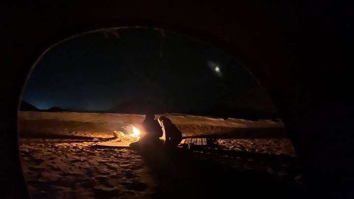 Silhouetted figures sit near a camp-fire viewed from inside a dark tent under starry sky