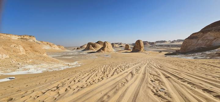 Tracks in golden sand lead toward clustered desert pinnacles beneath clear blue sky