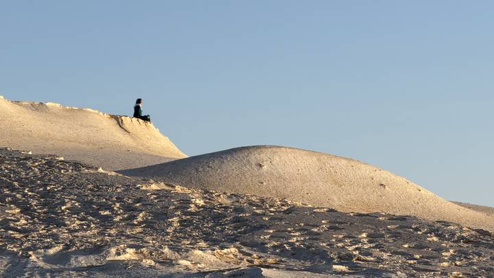 Traveller sits alone atop pale dune overlooking undulating desert landscape