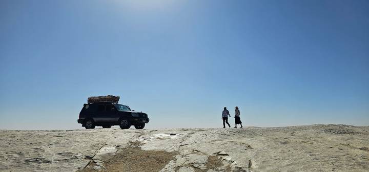 SUV and two travellers stand on bright rock plateau under cloudless sky
