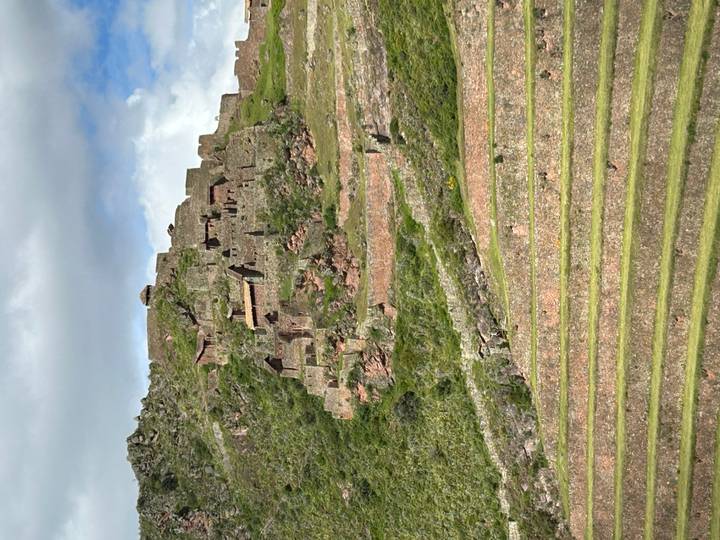 Ancient agricultural terraces climb a green hillside towards the stone ruins of Pisac under a cloudy sky.