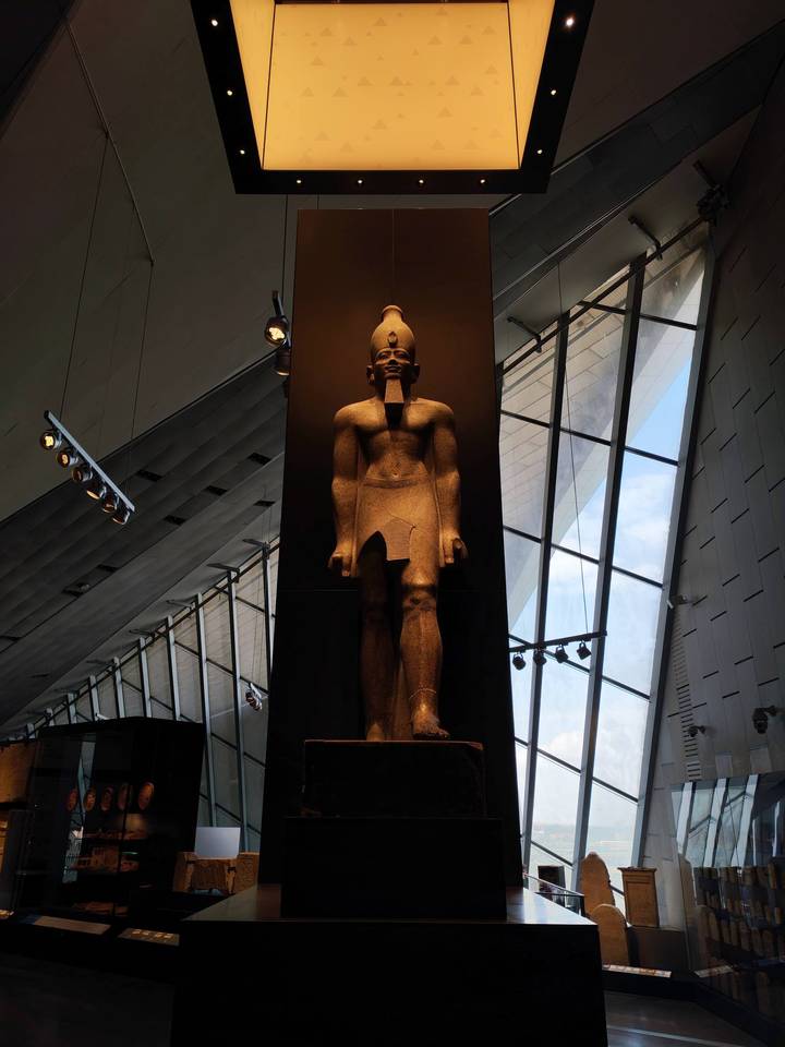 Colossal pharaoh statue displayed beneath the modern angular ceiling of the Grand Egyptian Museum.
