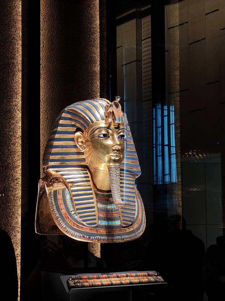 Gold and lapis funerary mask of Tutankhamun gleams under museum spotlights with reflections on glass.