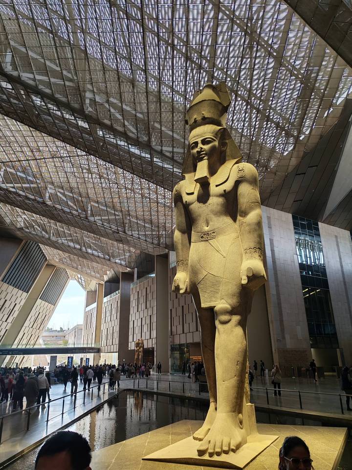 Massive sandstone pharaoh statue stands beneath a dramatic steel-and-glass roof of the museum atrium.