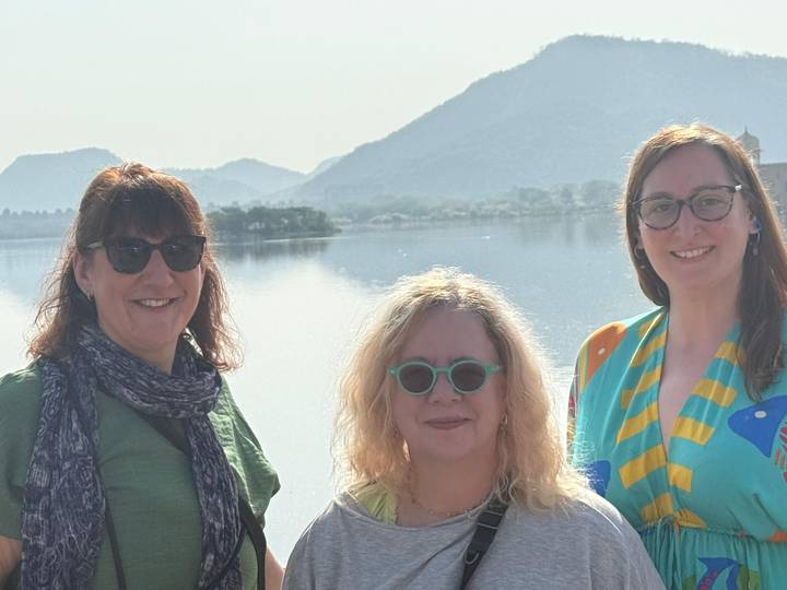 Three female travellers pose beside a tranquil lake with hazy Rajasthan hills in the background.