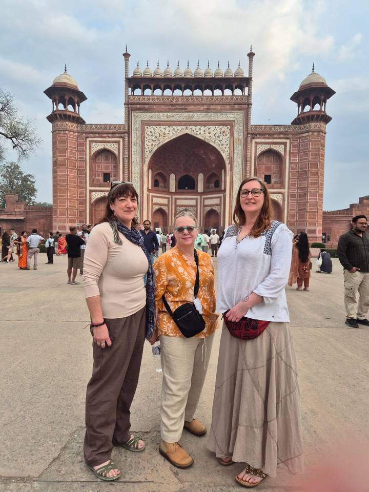 Three women pose in front of the grand red sandstone gateway of Agra Fort with crowds behind.