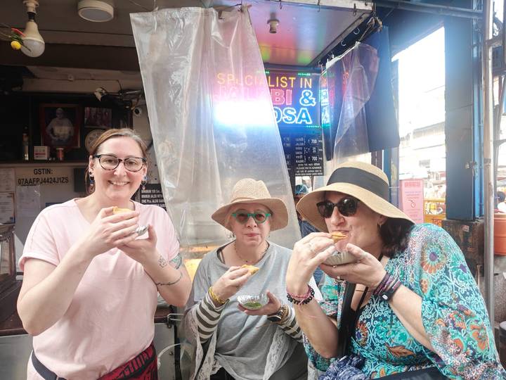 Women sample street snacks at a busy Delhi food stall lit by a neon sign.