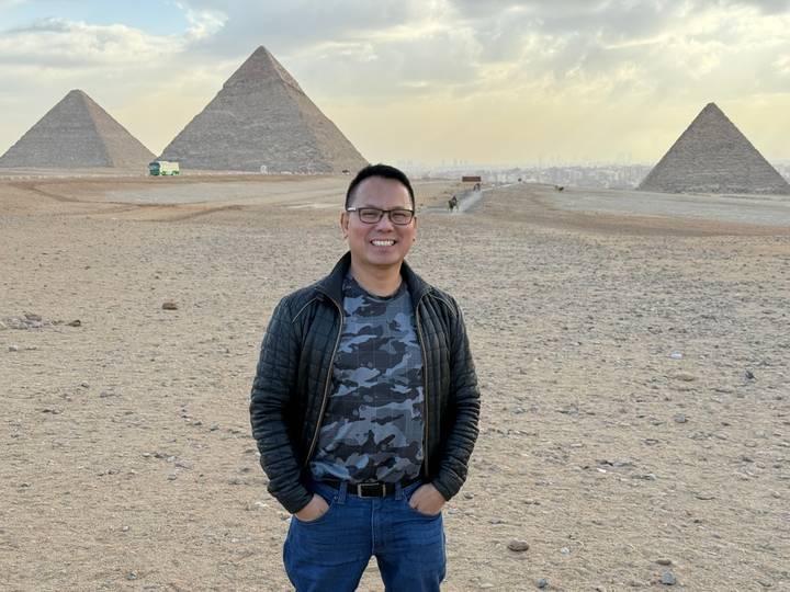 Traveler poses smiling with the three Pyramids of Giza rising behind him.