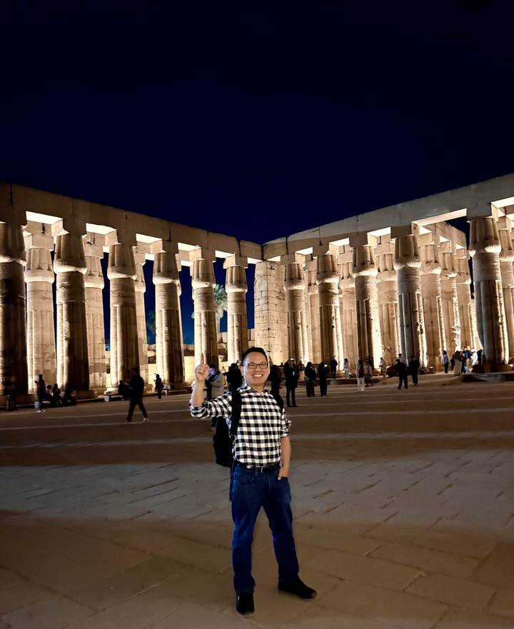 Visitor stands in front of illuminated columns of Luxor Temple at night.