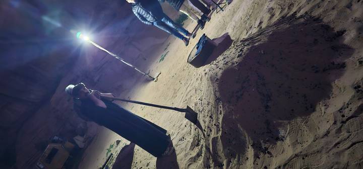 Bedouin guide uncovers a sand-buried meal beside guests under a bright lamp in the desert at night.