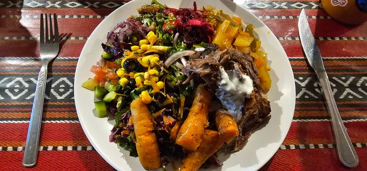 Colourful Middle-Eastern plate with salads, corn, roasted vegetables and slow-cooked meat on patterned tablecloth.