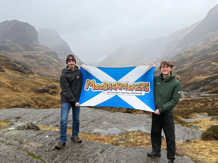 Two travellers hold a MacBackpackers flag amid the misty rugged mountains of Glencoe.