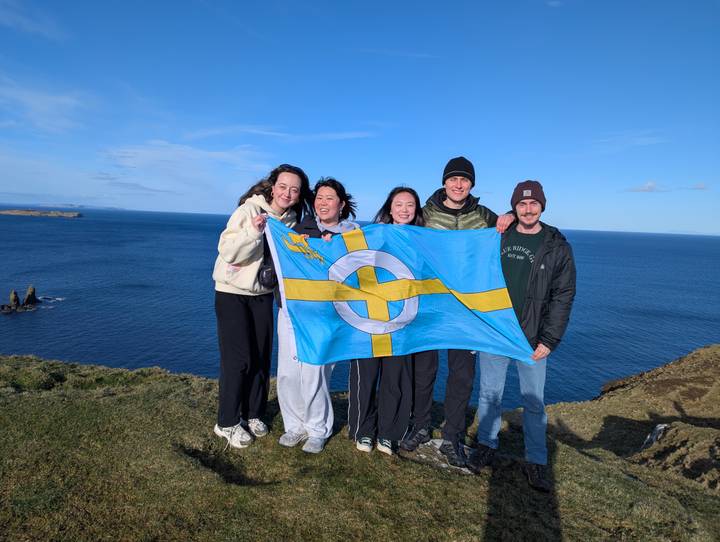 Travellers pose with a blue flag on a windy cliff edge above the vast Atlantic Ocean.