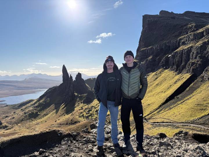 Two hikers stand beneath the jagged rock pinnacles of the Old Man of Storr on a sunny day.