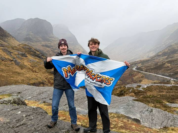 Two friends display a MacBackpackers flag in the dramatic, cloud-shrouded valley of Glencoe.