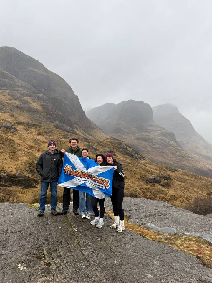 Group of travellers hold a MacBackpackers flag in front of looming Glencoe peaks under misty skies.