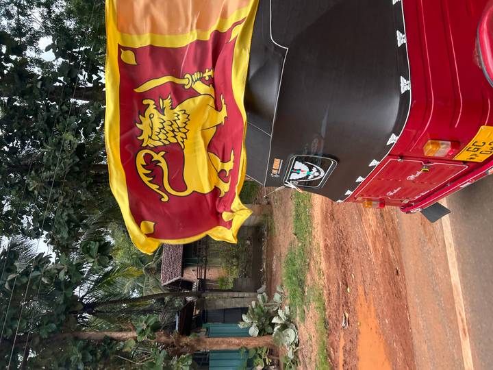 Red three-wheeled tuk-tuk adorned with a lion flag driving along a rural Sri Lankan road