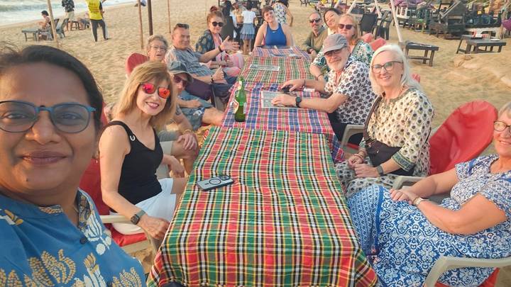 Tour group seated at long table on sandy beach enjoying sunset drinks