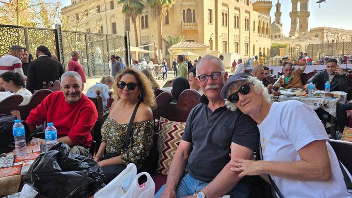 Four travelers relaxing at an outdoor cafe in historic Cairo with ornate buildings and mosque minarets in the background.