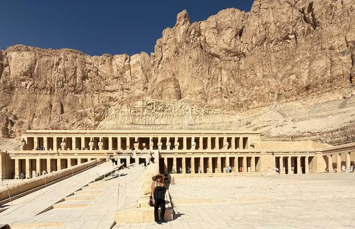 Temple of Hatshepsut carved into towering cliffs with a lone visitor in the vast forecourt under deep blue sky.