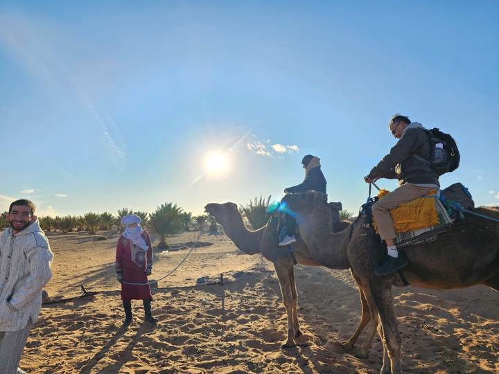 Travelers and guides stand with camels in the desert as the low afternoon sun creates a warm glow.