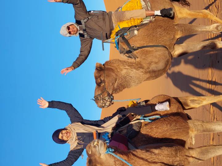 Smiling riders wave while seated on camels against tall orange dunes and clear blue sky.