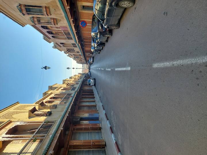 Empty straight street lined with traditional Moroccan buildings and shuttered shops under a clear morning sky.