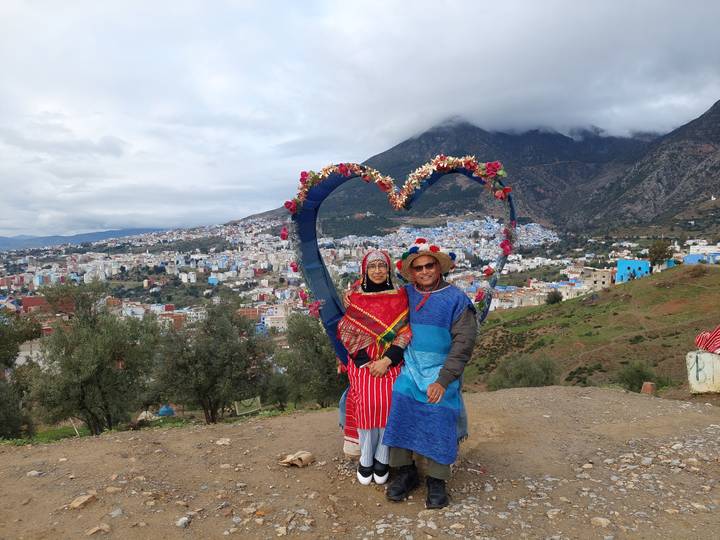 Couple in colorful attire stands before a heart-shaped frame overlooking the blue hillside town of Chefchaouen and surrounding mountains.