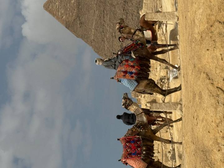 Locals ride decorated camels with a pyramid silhouette against a partly cloudy sky.