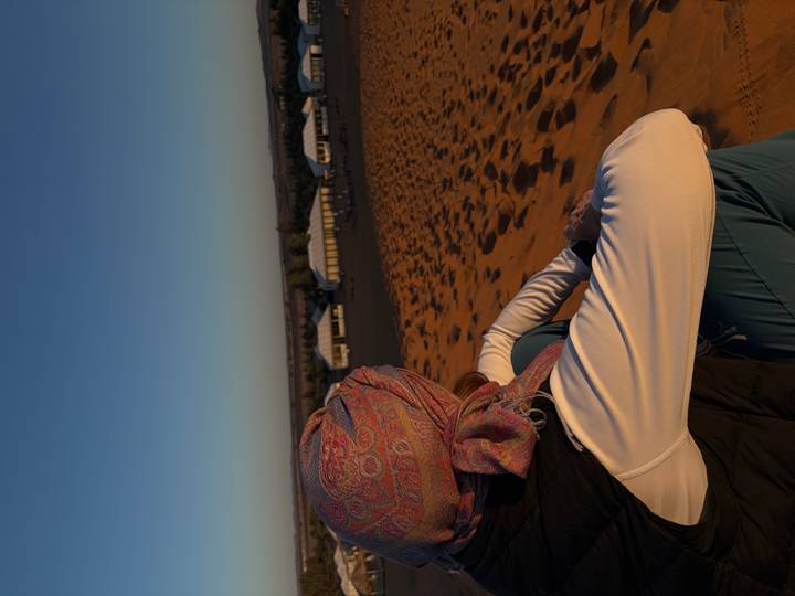 Traveler in headscarf sits on a dune at dawn overlooking a desert tent camp.