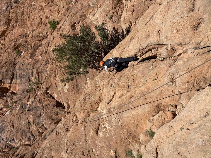 Climber wearing helmet ascends a steep rock face with ropes in a canyon.