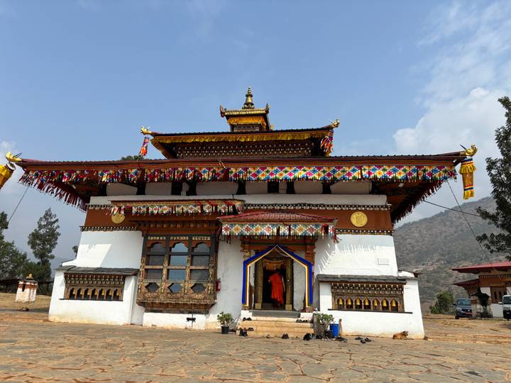 Whitewashed Bhutanese temple with brightly colored tiered roof, golden ornaments and prayer flags set against a hazy mountain backdrop.