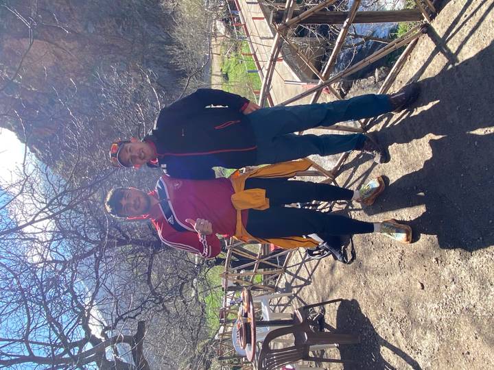 Two hikers pose on a sunny mountain trail with bare trees and rocky peaks in the Atlas Mountains.