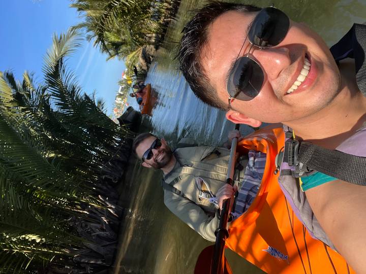 Two friends smile in orange kayaks paddling through a palm-lined waterway on a sunny day.