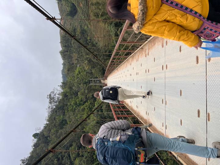 Travelers cross a long metal suspension bridge high above a forested valley on a cloudy day.