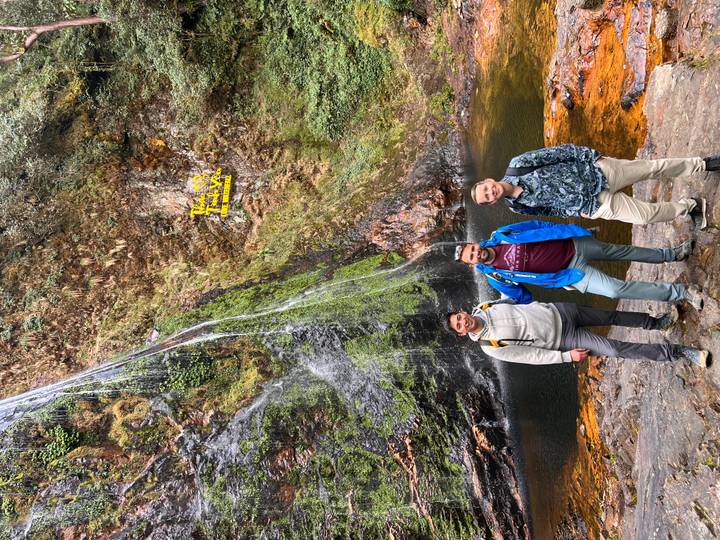 Three travellers pose cheerfully beside a forest waterfall cascading into a dark pool