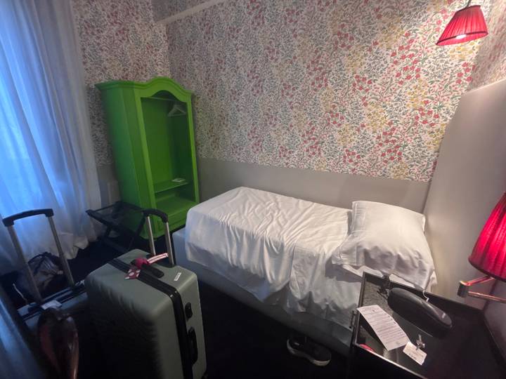 Small single hotel room with floral wallpaper, green wardrobe and luggage