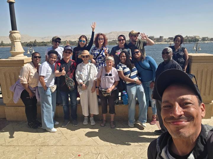 Large tour group smiling on a riverside promenade with desert hills across the water.