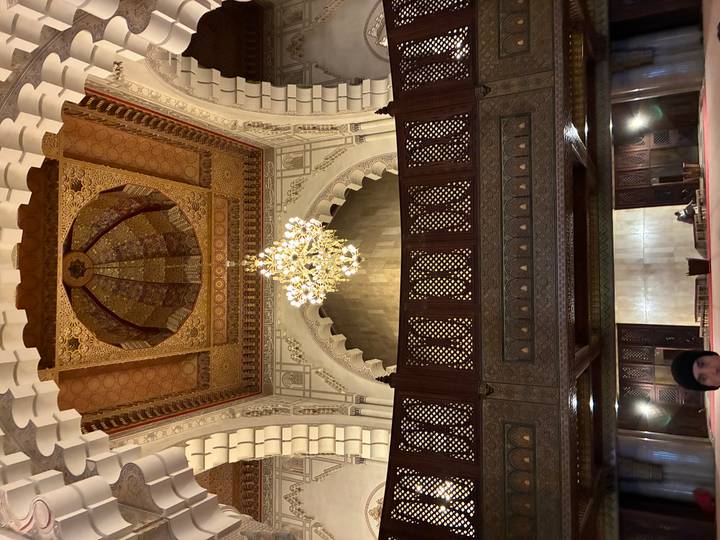 Lavishly decorated mosque interior with grand chandelier and carved wooden gallery.