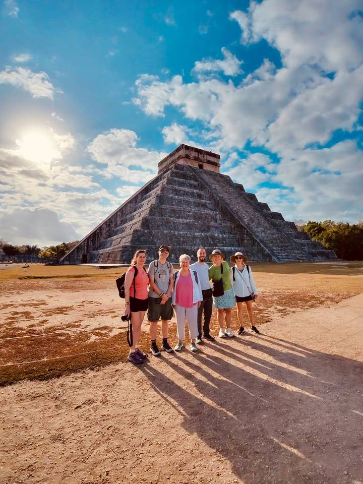 Travel group poses before the iconic pyramid of Chichen Itza under a partly cloudy sky.